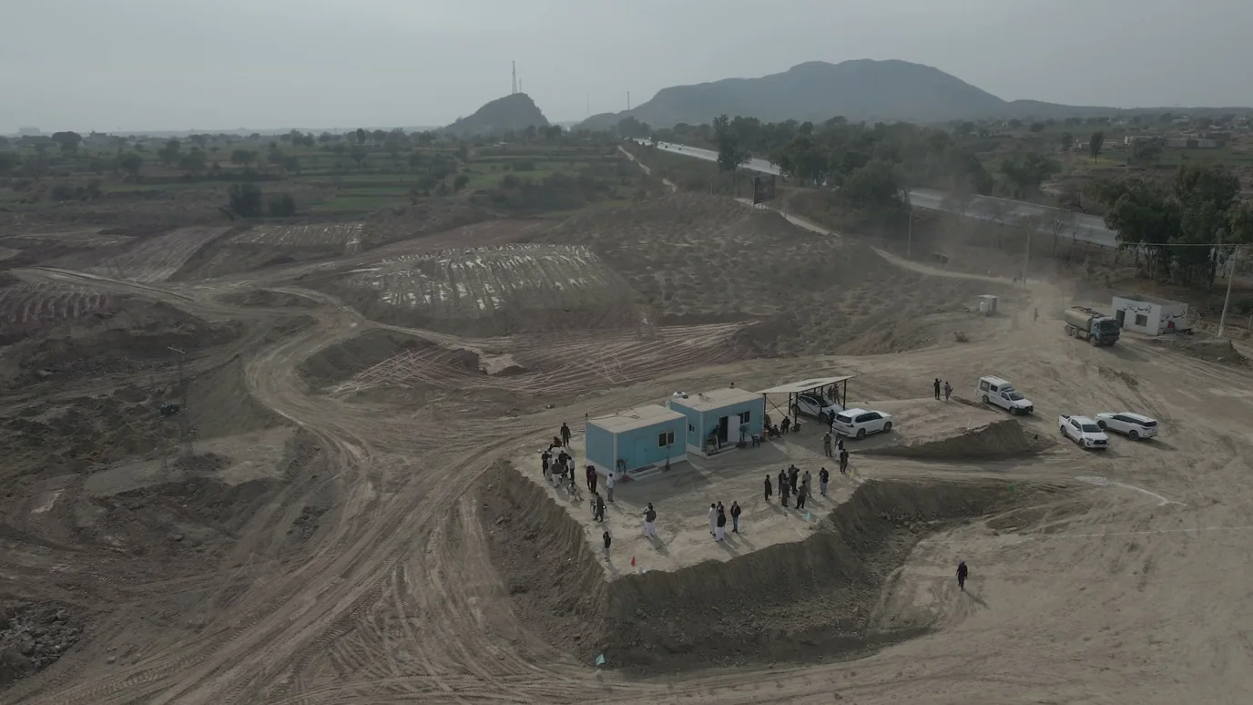 Drone view of bare graded land with a site office of container cabins, parked cars and a crowd, a tree-lined highway and hills beyond