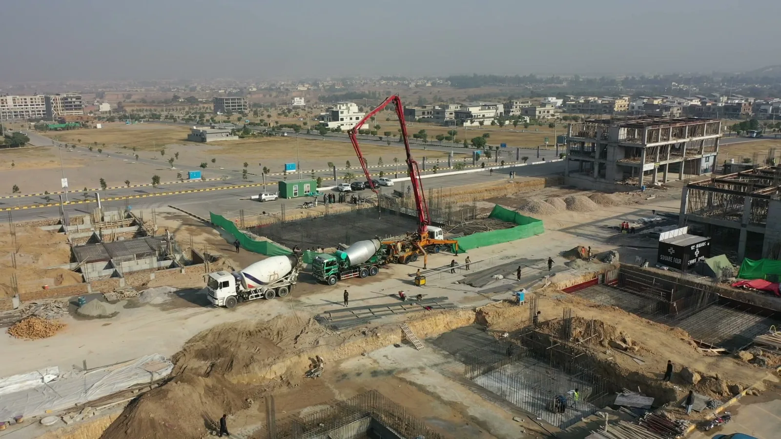 A drone view of a basement slab being poured on a Pakistani construction site, with steel reinforcement and footings in the open excavation
